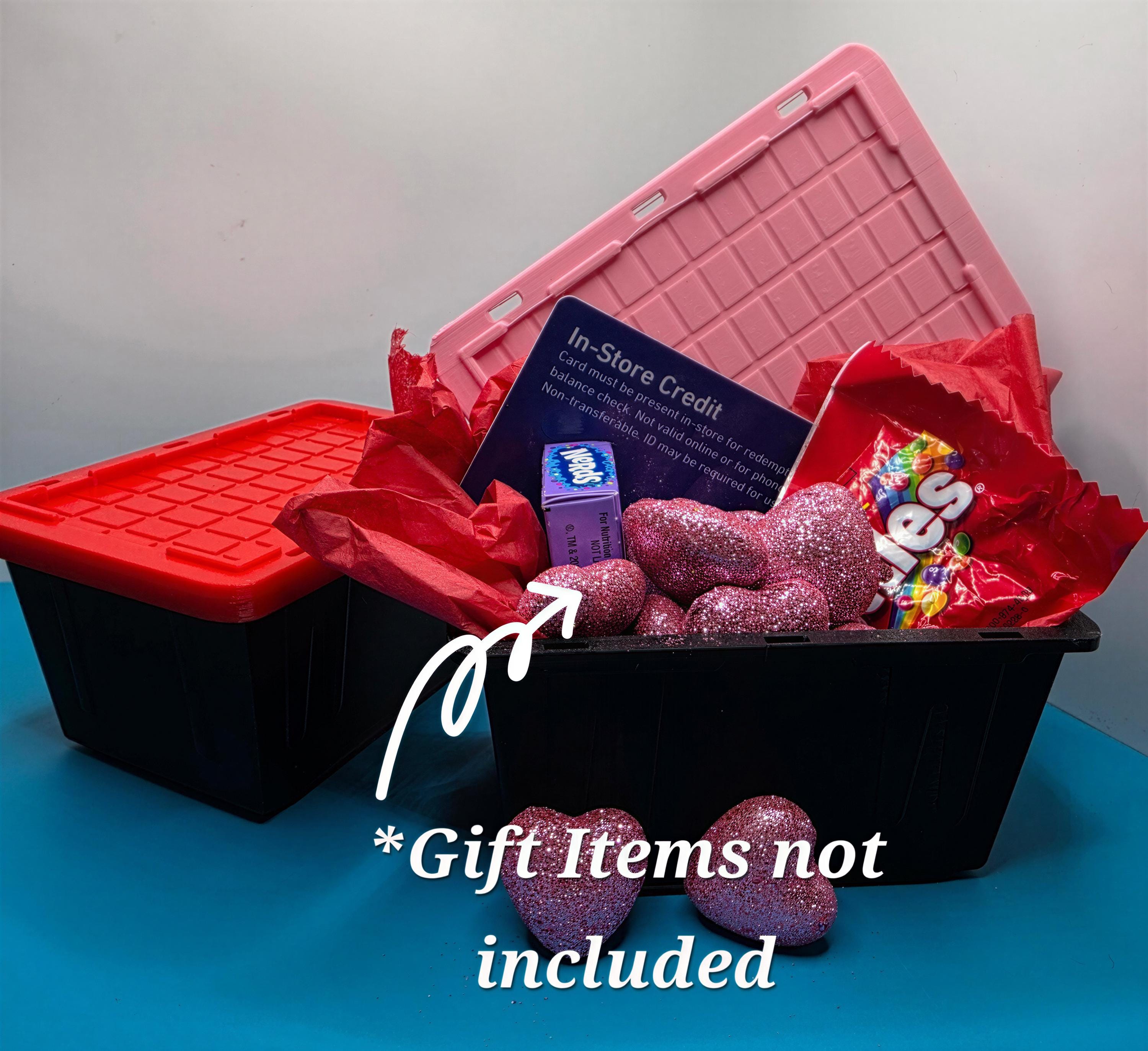 A gift basket containing various items, including a pink plastic container, a red plastic container, and a pink glittery item, along with a card and a small blue item. The basket is placed on a blue surface, and there is a text overlay indicating that the gift items are not included.