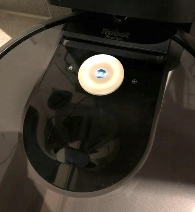 May include: Close-up view of a Roomba robot vacuum's charging dock. A circular, off-white charging sensor is prominently displayed on the dock's surface. The Roomba logo is visible on the top of the dock. The dock is black and glossy.  The home base icon is visible next to the sensor.