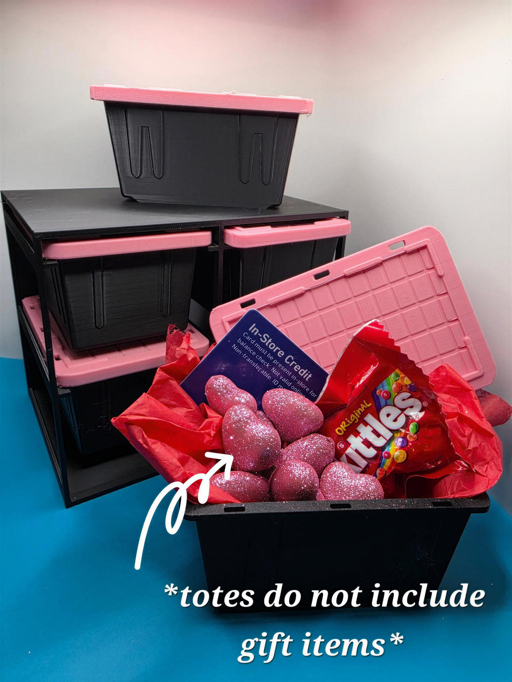 A pink storage bin filled with various items, including a bag of candy, a pink bag, and some pink glittery objects, with a black storage bin and pink storage bin stacked on top.